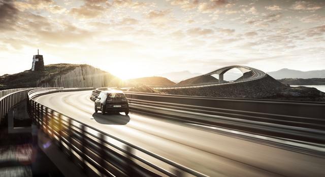 Car driving on a winding, elevated roadway, with a bridge, mountains, hills, and a body of water in the distance